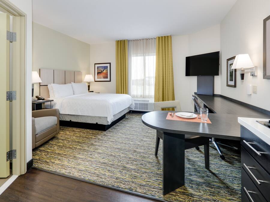 A hotel room with a bed, bedside table, lamp, TV, and a desk with a chair.