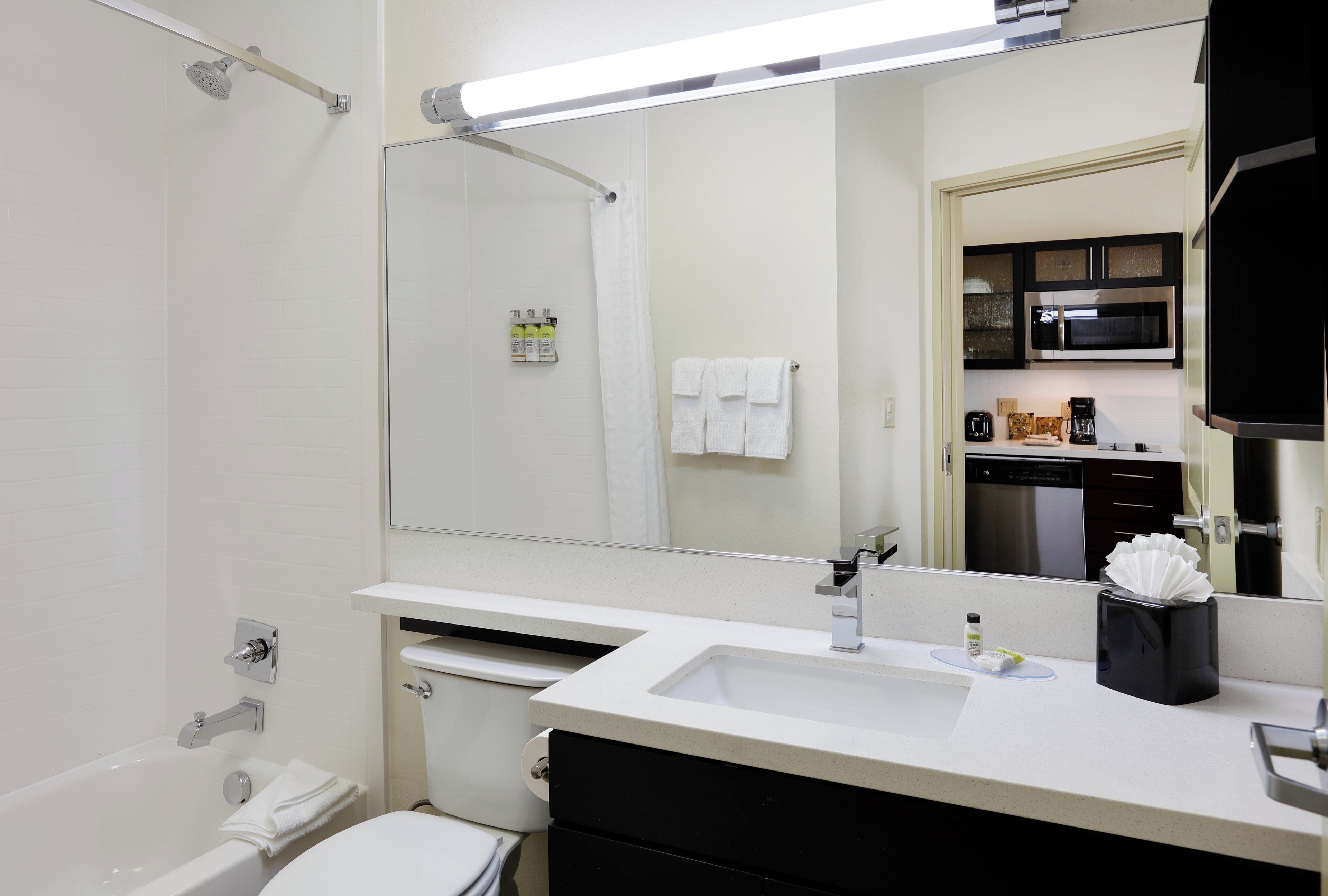 A modern bathroom with a bathtub, toilet, sink, and a large mirror reflecting a kitchen area.