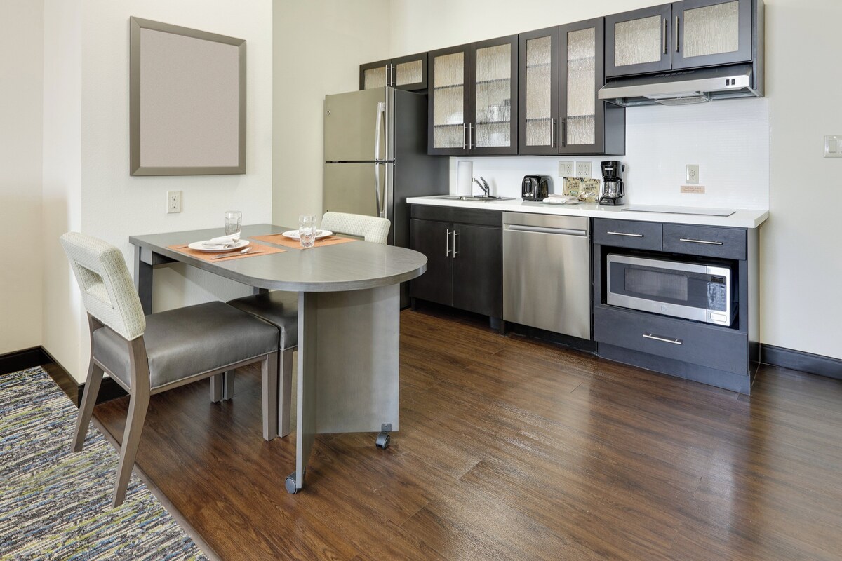 Cozinha moderna com eletrodomésticos de aço inoxidável, sala de jantar com mesa e cadeiras, e tapete estampado no chão.