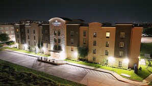 A multi-story hotel with a stone facade, a well-lit entrance, and a parking lot. - Candlewood Suites Dallas NW - Farmers Branch by IHG (Farmers Branch)
