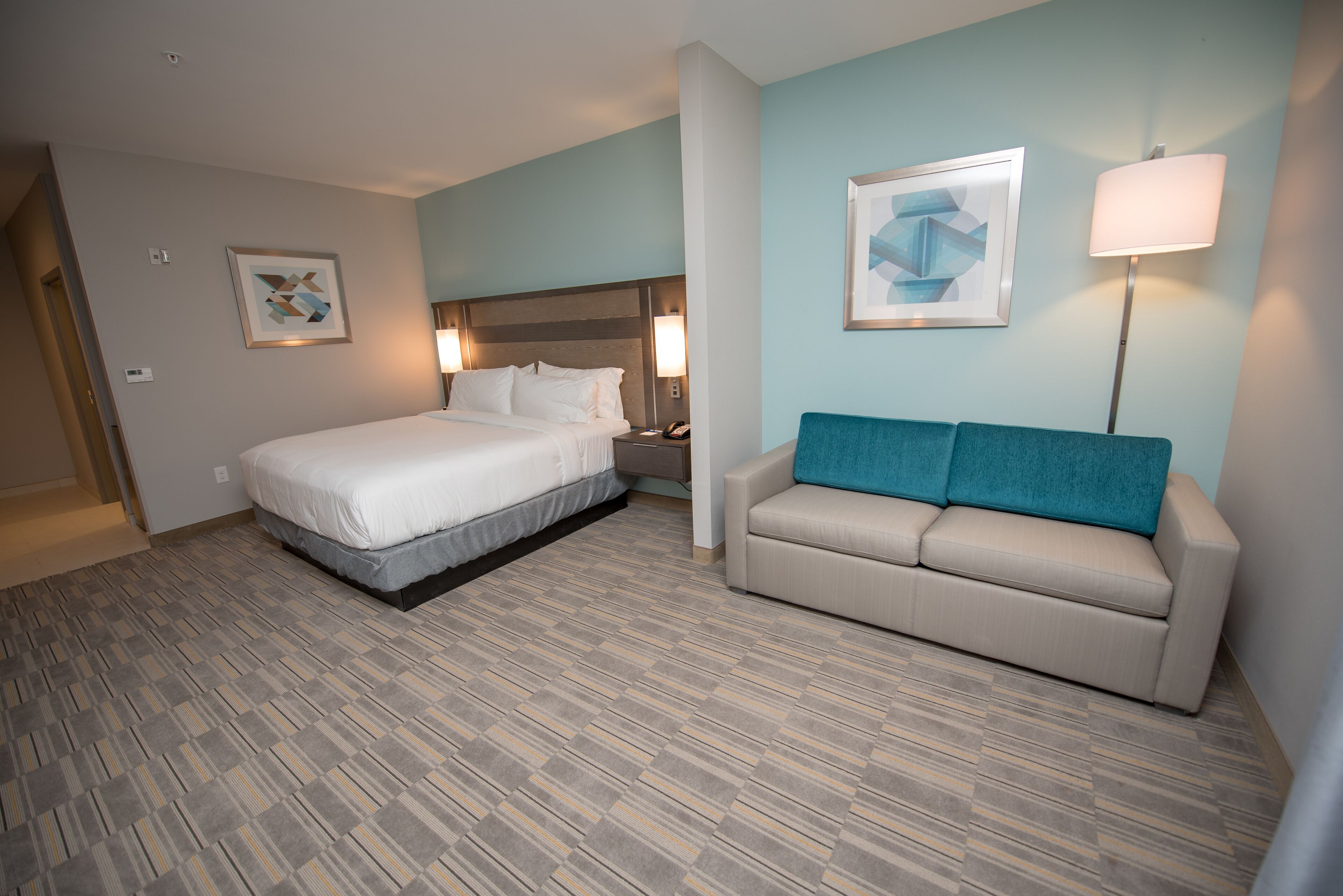A hotel room with a bed, a sofa, a nightstand, a lamp, and two framed pictures on the wall.