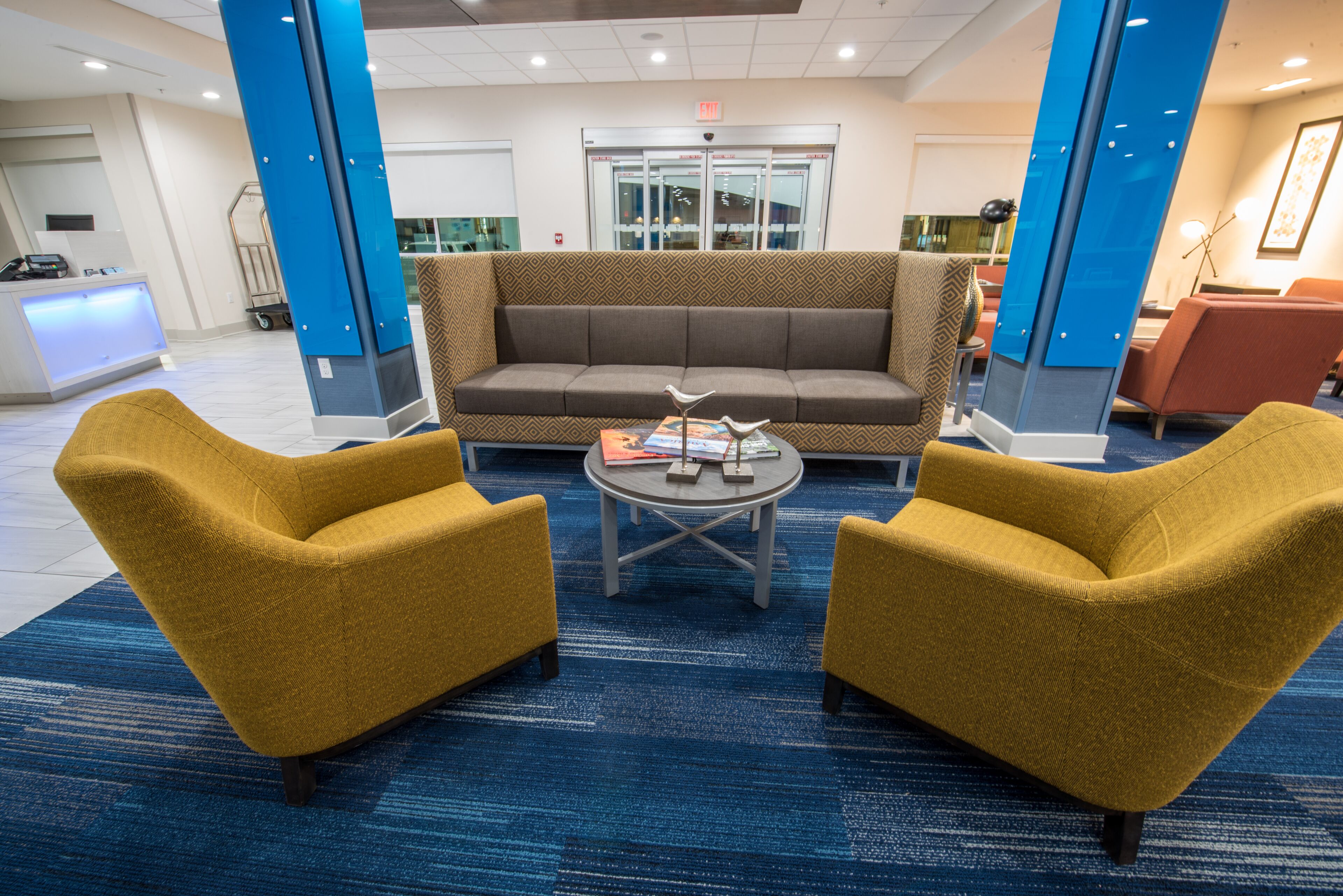 Um hall de entrada com um balcão de recepção, um sofá marrom, duas poltronas amarelas e uma mesa de centro com livros e uma escultura de pássaro.