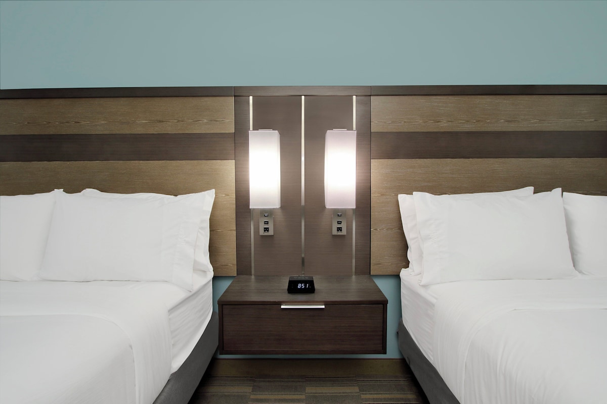 A hotel room with two beds, a nightstand with a digital clock, and wall-mounted lighting.
