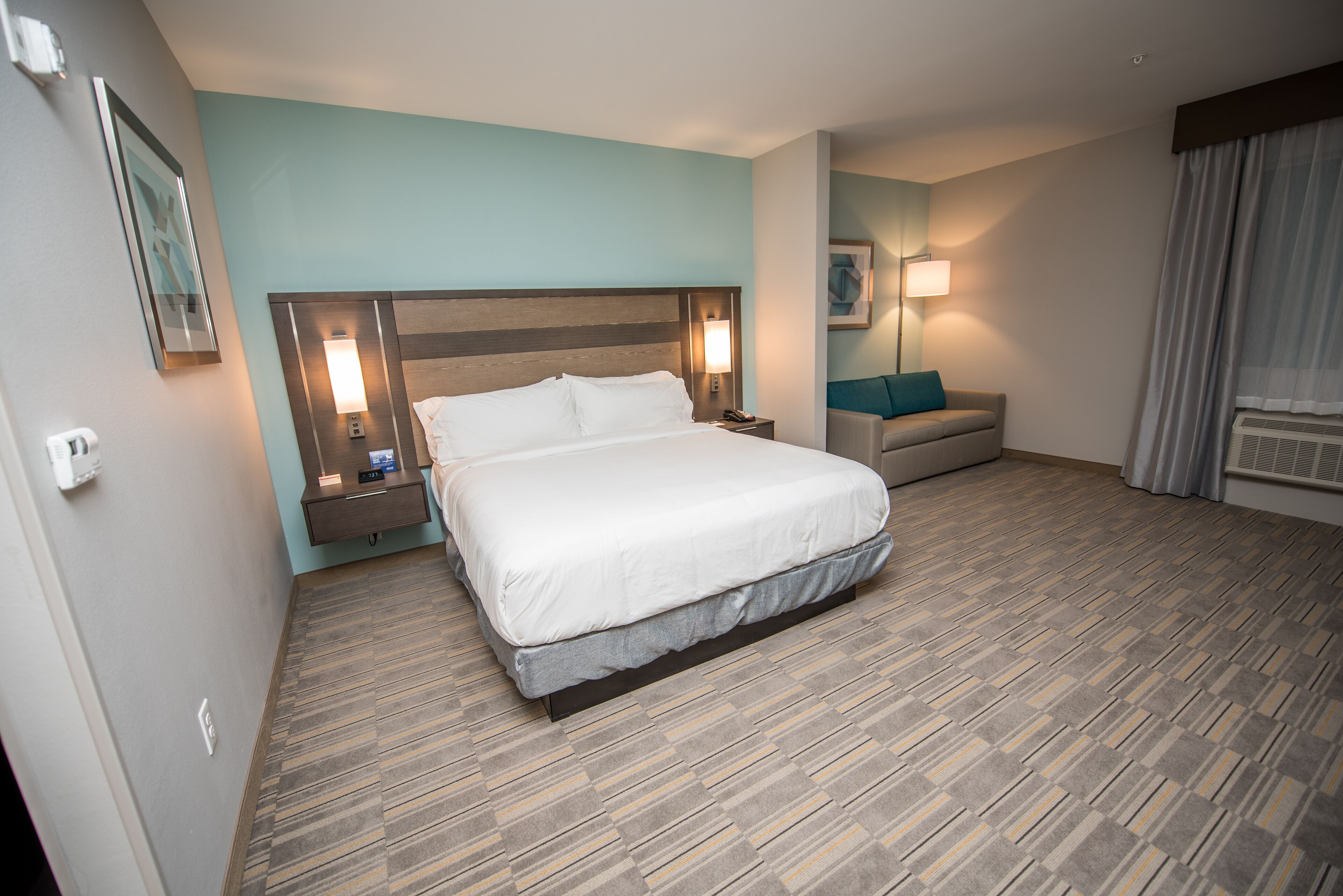 A hotel room with a large bed, a bedside table with a lamp, a sofa, and a wall-mounted TV.