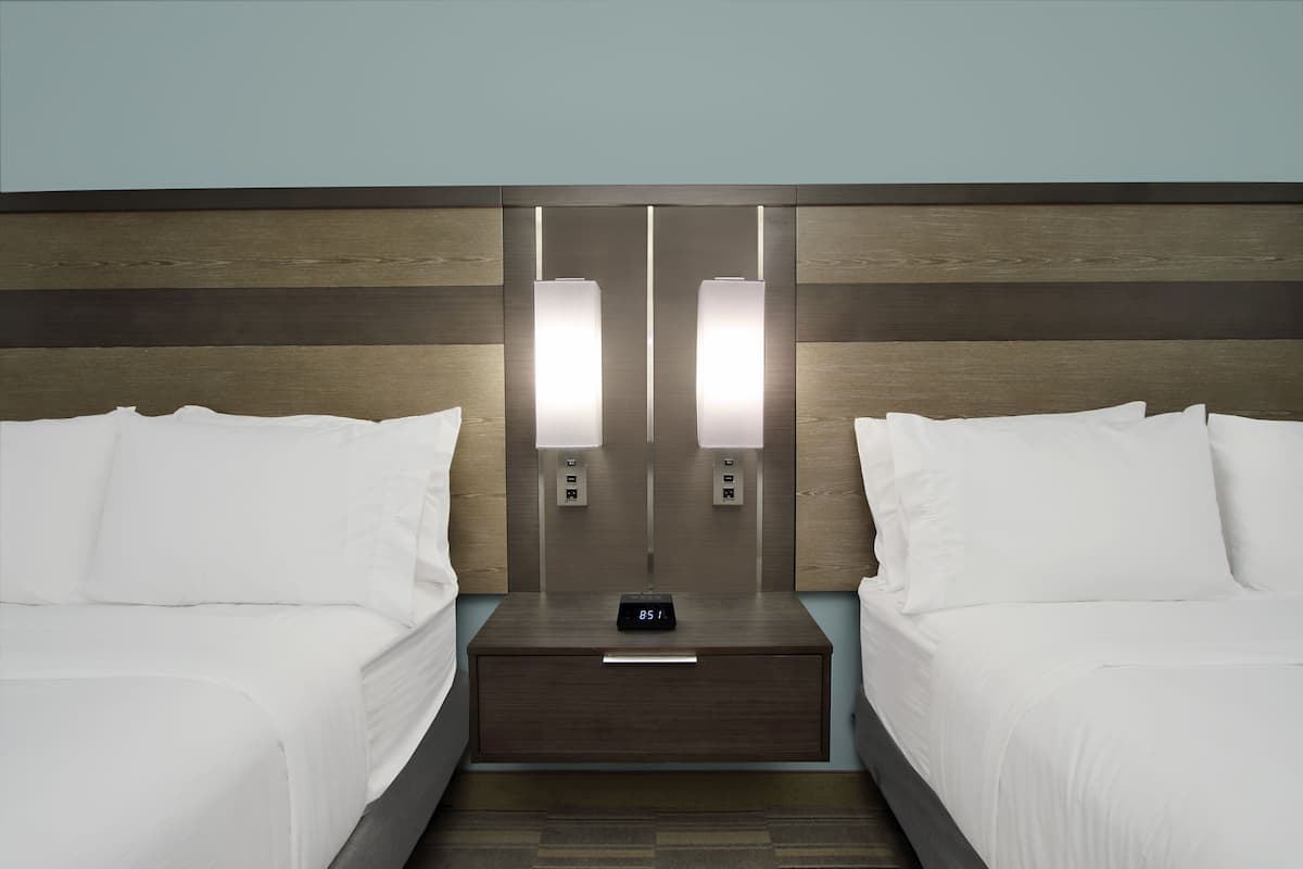 A hotel room with two beds, a nightstand with a digital clock, and wall-mounted lights.