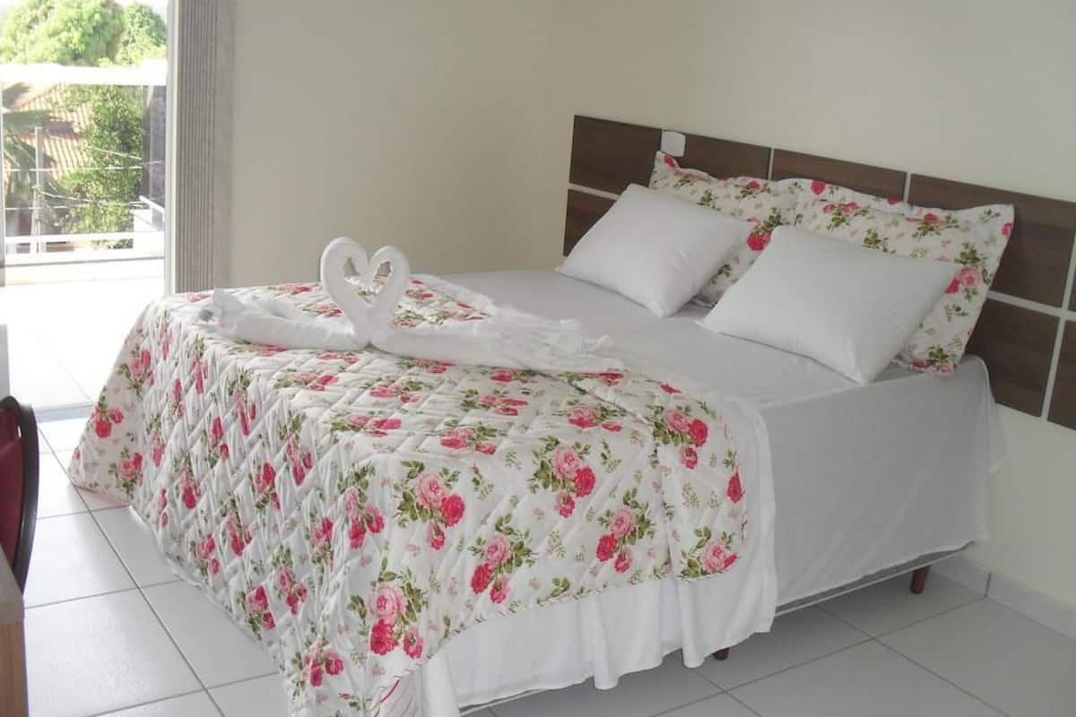 A neatly made bed with floral bedding and white pillows, a heart-shaped towel, and a view of the outdoors through a window.