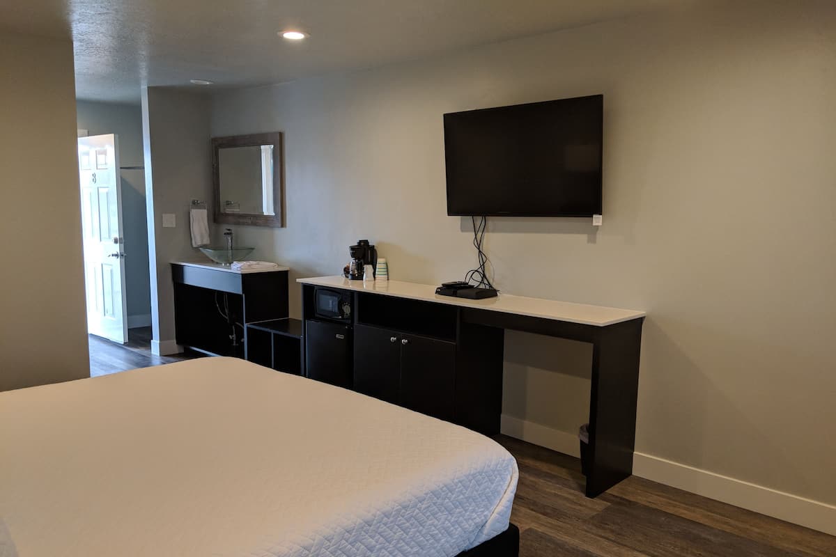 A hotel room with a bed, a desk with a flat-screen TV, a small kitchenette, and a mirror.