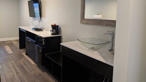 A modern bathroom with a glass sink, a coffee maker, and a flat-screen TV mounted on the wall.