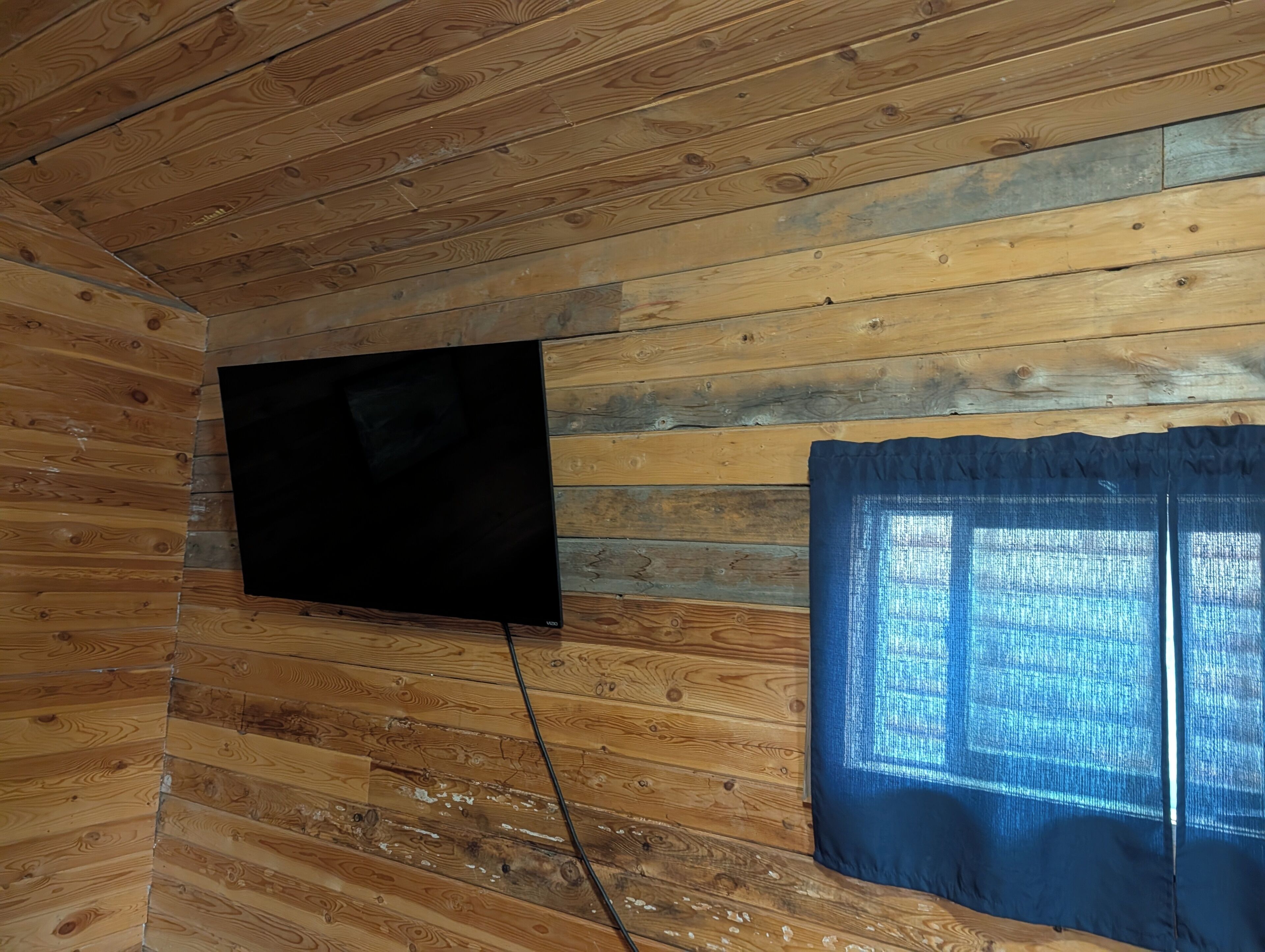 A room with wooden walls, a flat-screen TV mounted on the wall, and a window with blue curtains.