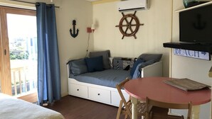 A bedroom with a bed, a small table, a chair, a ceiling fan, an air conditioning unit, a TV, and a window with curtains.