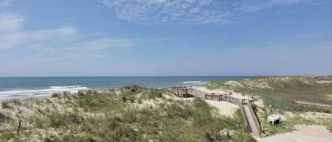 A beach with a wooden boardwalk leading to a sandy area, surrounded by dunes and vegetation.