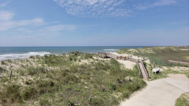 A beach with a wooden boardwalk leading to a sandy area, surrounded by dunes and vegetation.