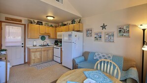 A compact kitchen with wooden cabinets, a refrigerator, and a microwave. There is a dining area with a table and chairs, and a sofa.
