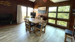 Een houten interieur met een eettafel, stoelen, een open haard en grote ramen.