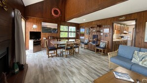 Een ruime woonkamer met houten vloer, een eettafel met stoelen, een bank en een keuken die zichtbaar is door een open doorgang.