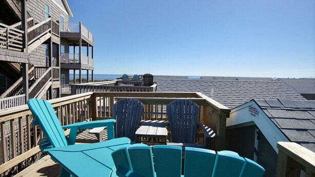 A wooden deck with blue Adirondack chairs and a table, overlooking the ocean. - 34 Sailing In A Gale Studio Bedroom Condo by RedAwning (Hatteras)