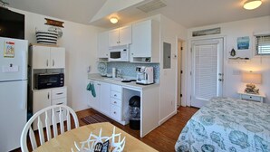A compact kitchen with a microwave, toaster, and coffee maker. There is a dining table with chairs and a bed with a patterned bedspread.