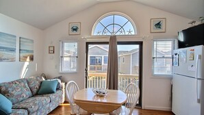 A cozy living room with a sofa, a dining table with chairs, a large window with a view, and a kitchen area with a refrigerator.