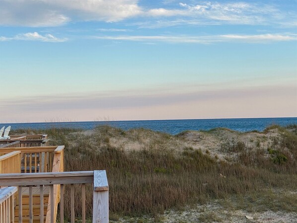 Terraza de madera con vistas a una playa, dunas y el océano al fondo.