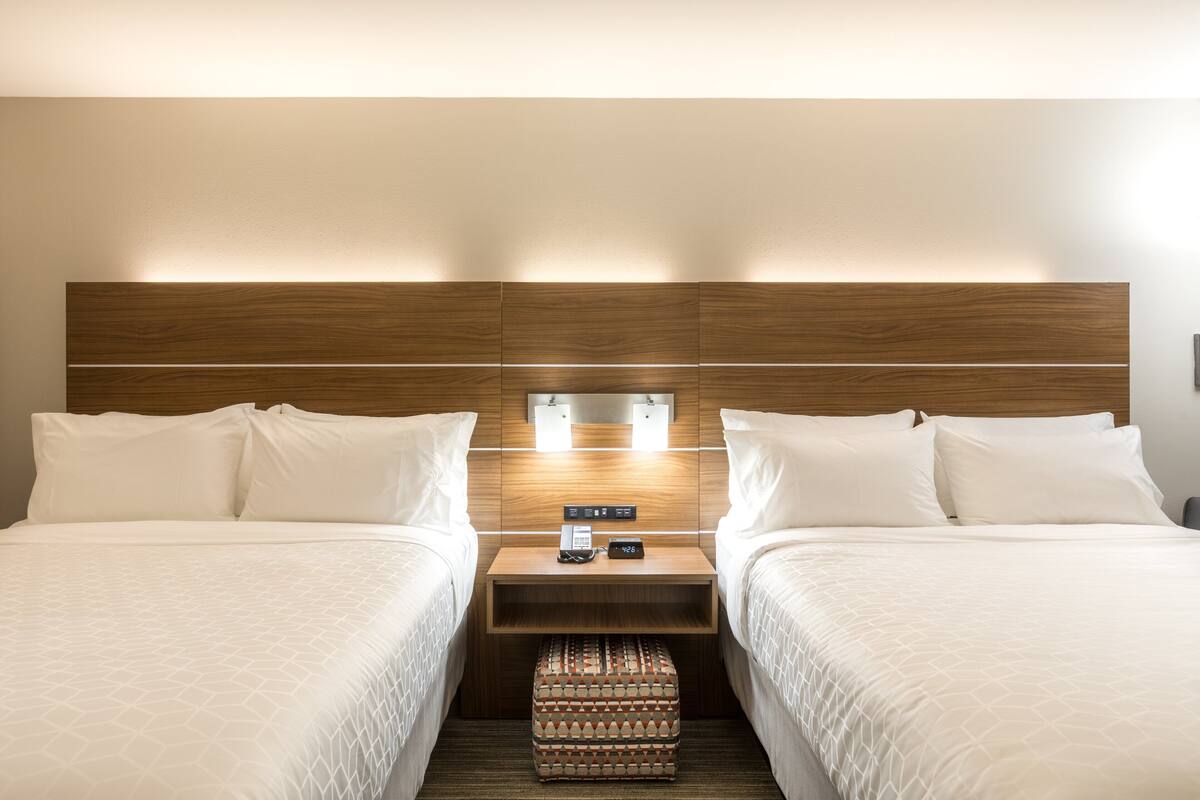 A hotel room with two beds, a wooden headboard, bedside lamps, a nightstand with a phone and a clock, and a patterned ottoman.