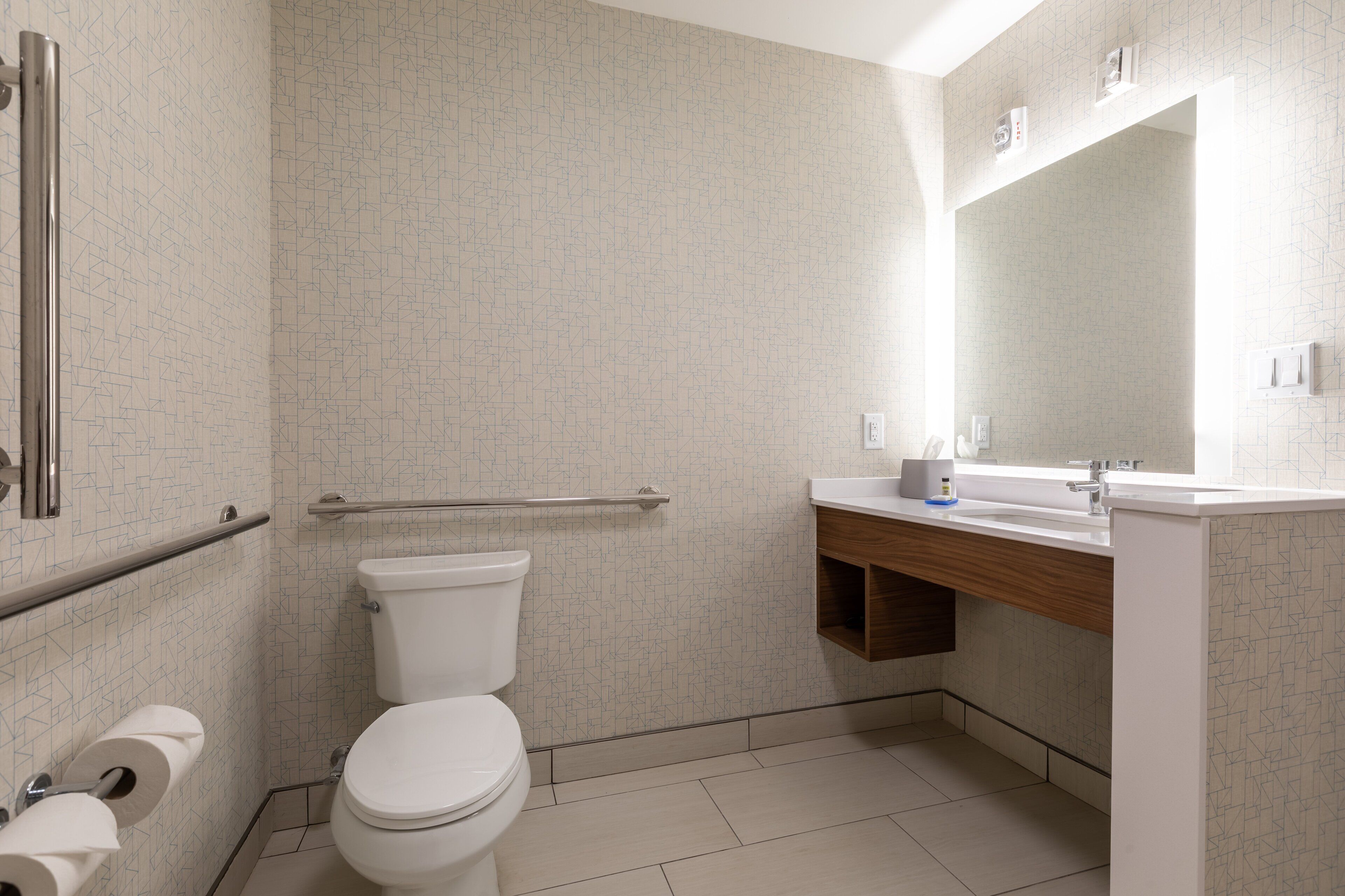 A modern bathroom with a toilet, handrail, sink, mirror, and toilet paper.