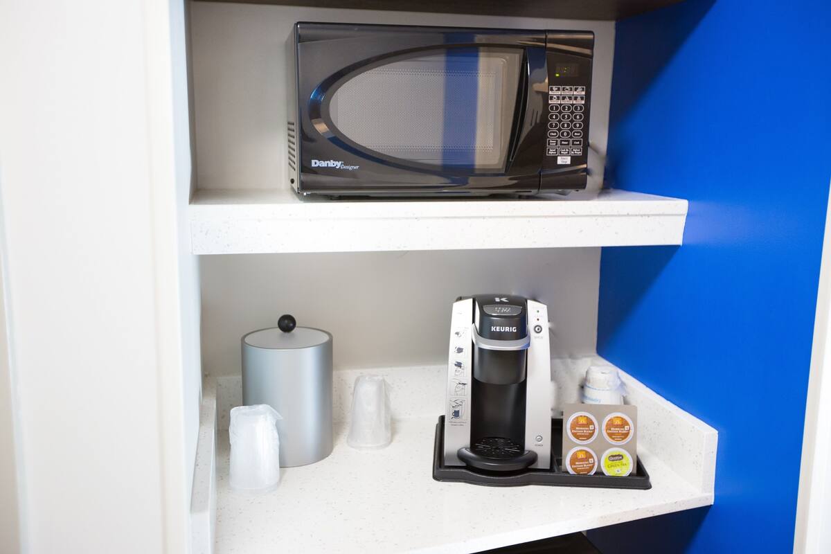 A kitchenette with a microwave, coffee maker, and coffee pods on a white countertop.