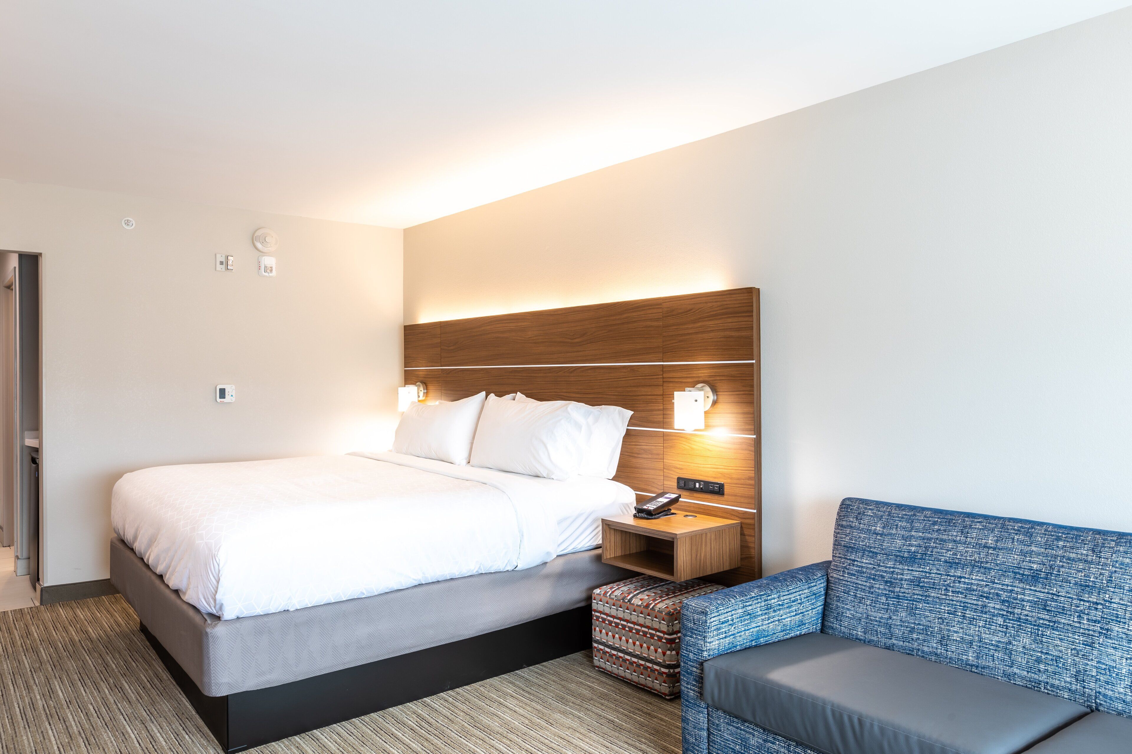 Una habitación de hotel moderna con una cama bien hecha, una mesita de noche con lámpara y un sofá azul.