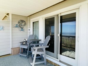 A covered outdoor area with a table and chairs, a decorative wall piece, and a view through a sliding glass door. - Sw 4 Seas-the-day 1 Bedroom Condo by RedAwning (Hatteras)