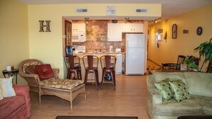 Ein Wohnzimmer mit einer Couch, einem Sessel, einem Esstisch mit Stühlen, einer Küchenzeile mit Schränken und Geräten sowie einer Pflanze.