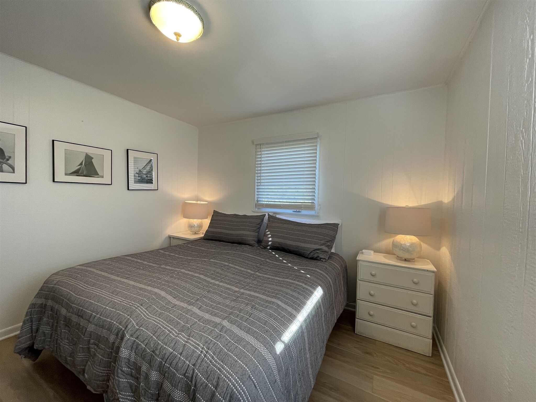 A bedroom with a bed, bedside lamps, a nightstand, a window with blinds, and framed pictures on the wall.