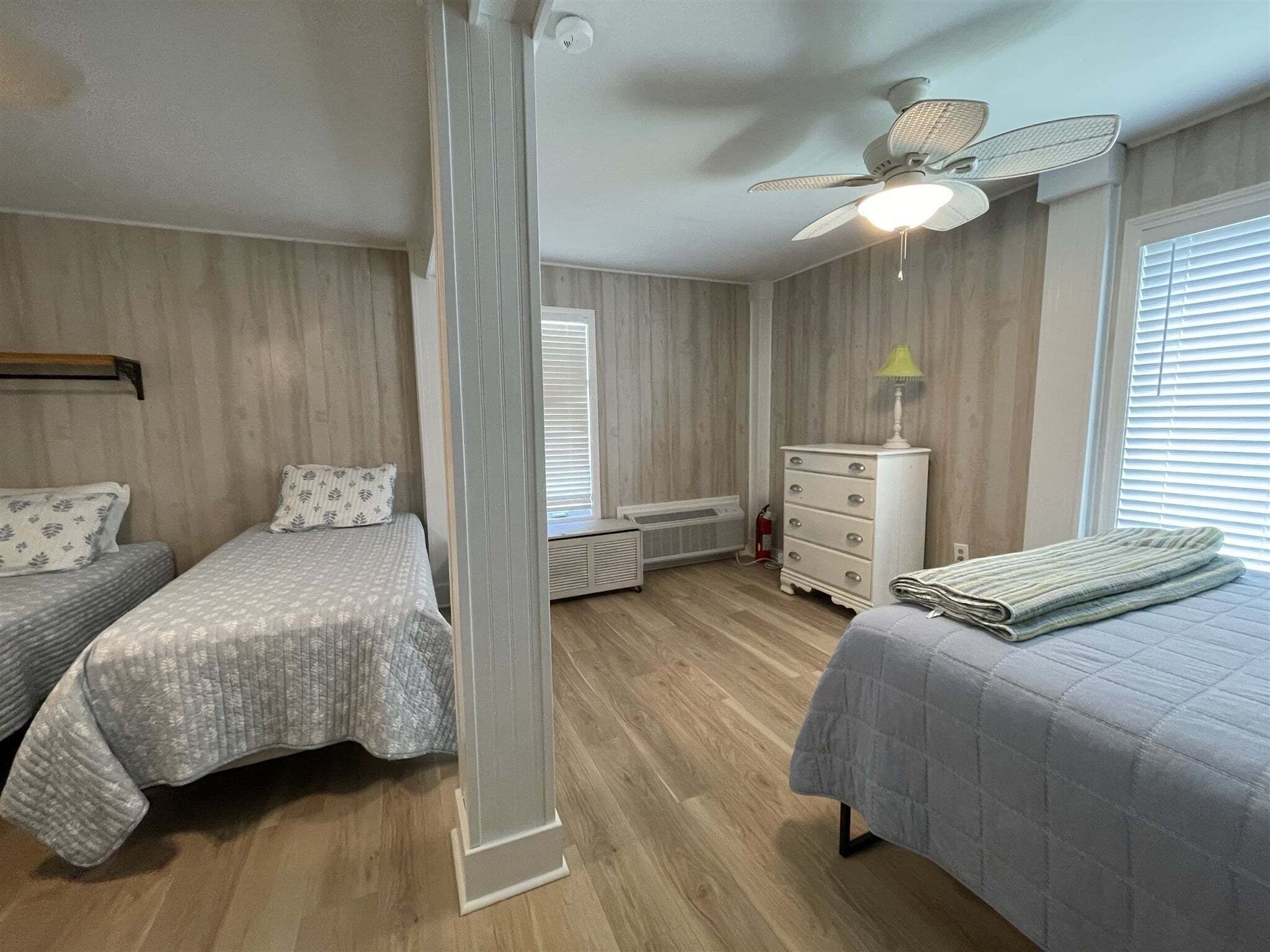 Une chambre avec deux lits simples, une commode, un radiateur et un ventilateur de plafond.