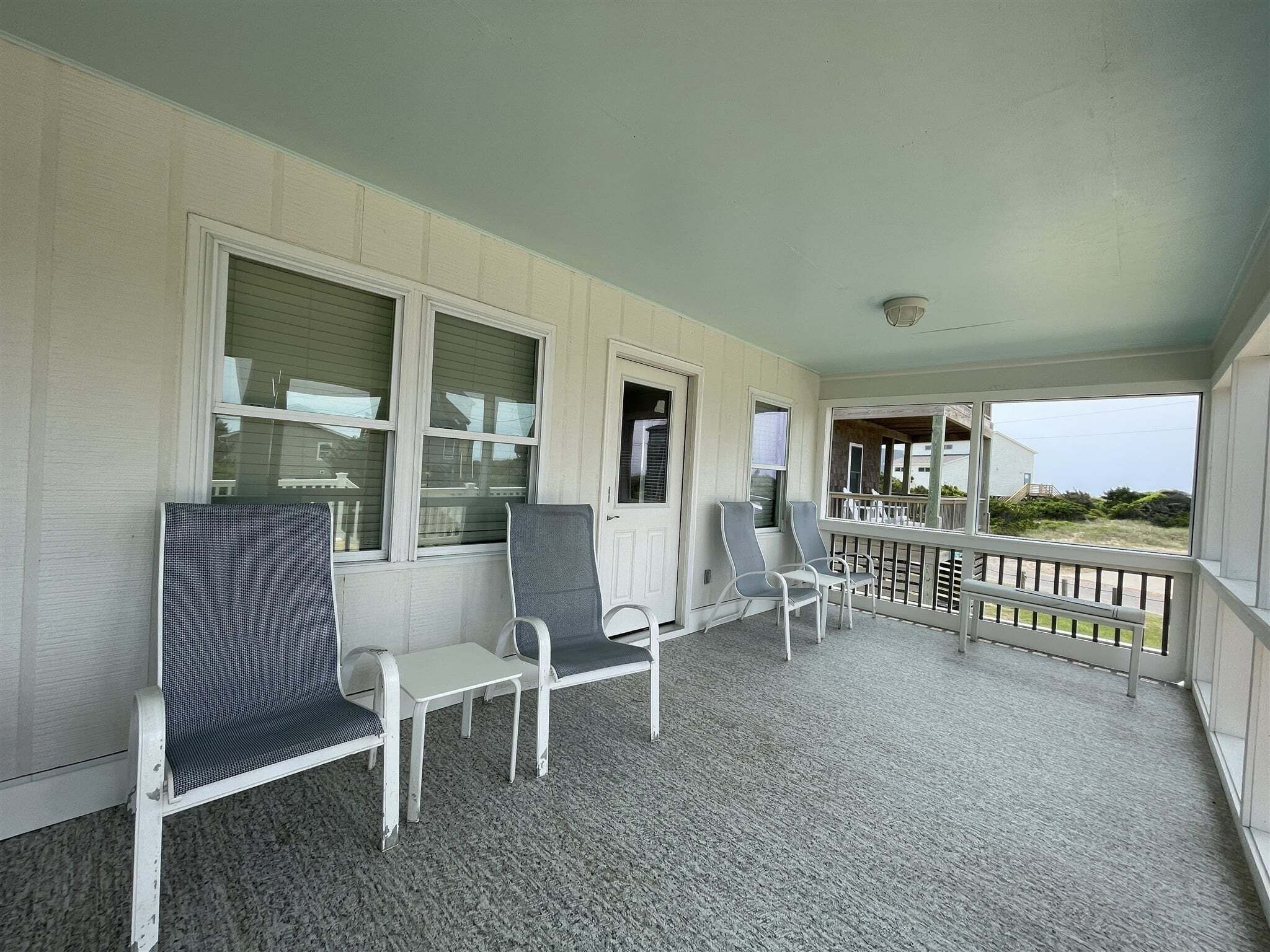 A covered porch with white chairs and a table, a door, and a view of the outside.