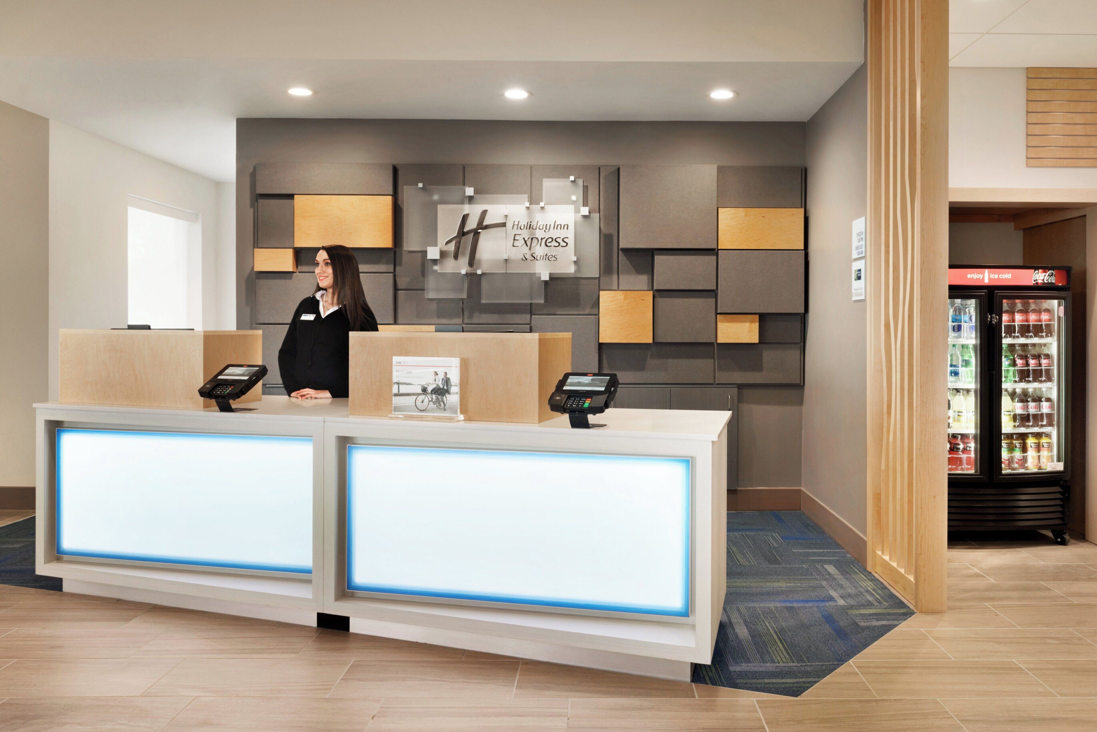 A hotel reception desk with a digital display, a Coca-Cola refrigerator, and a wall-mounted sign for Holiday Inn Express & Suites.