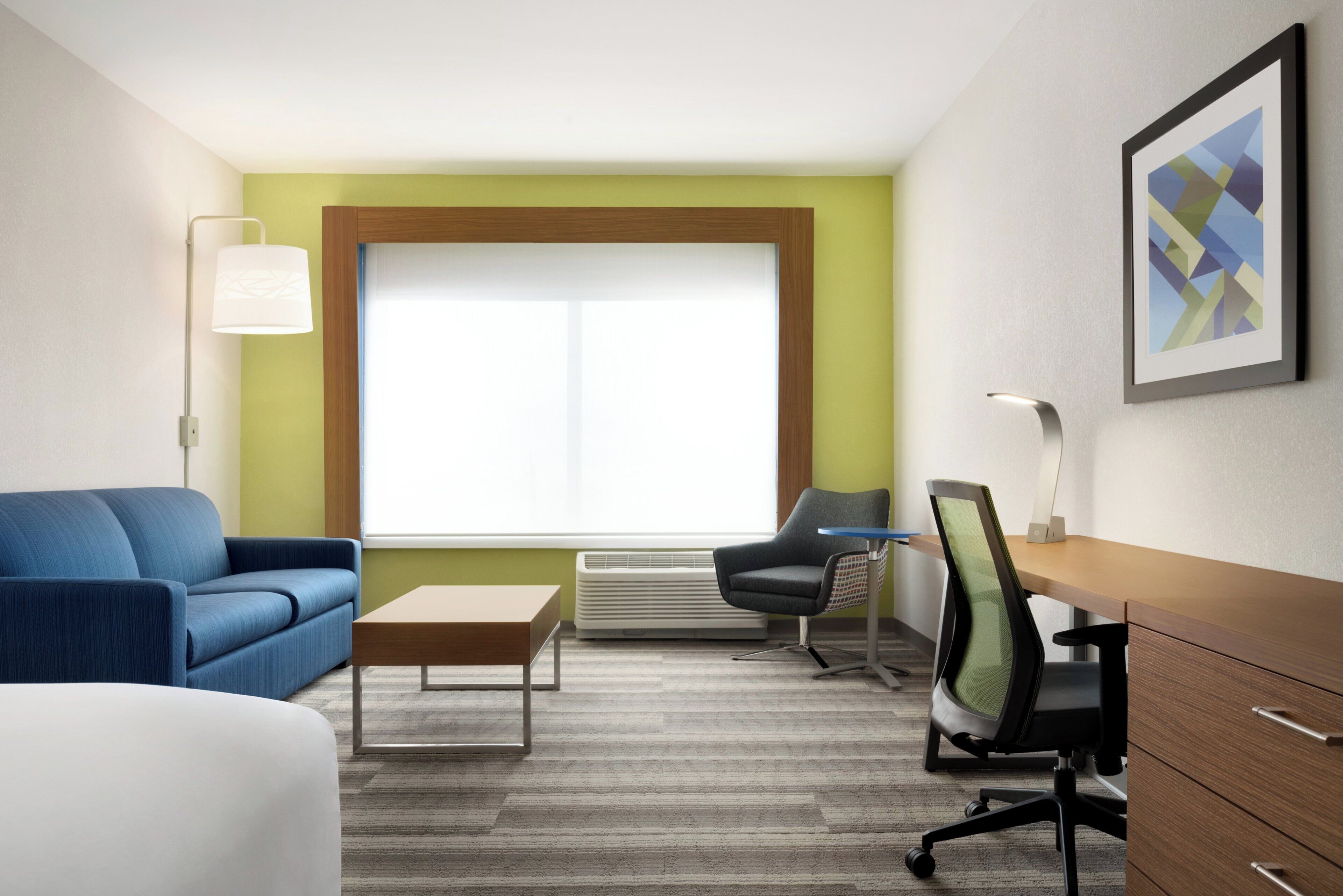 A hotel room with a blue sofa, a wooden desk, a chair, a lamp, and a large window.