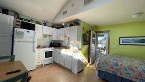 A compact kitchen with white cabinets, a stove, and a microwave. There's a dining table with chairs and a bed with a floral bedspread.