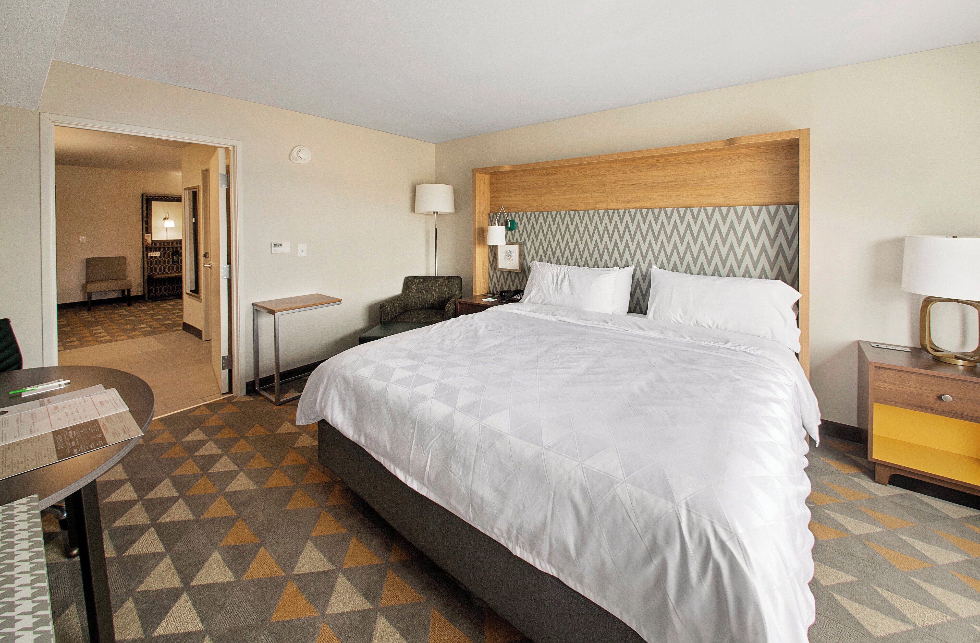 A hotel room with a large bed, a desk, a chair, and a nightstand with a lamp.