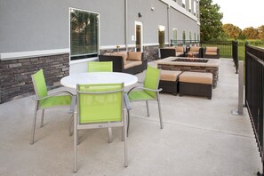En udendørs terrasse med et rundt bord og stole, en bålplads og et siddeområde med hynder.