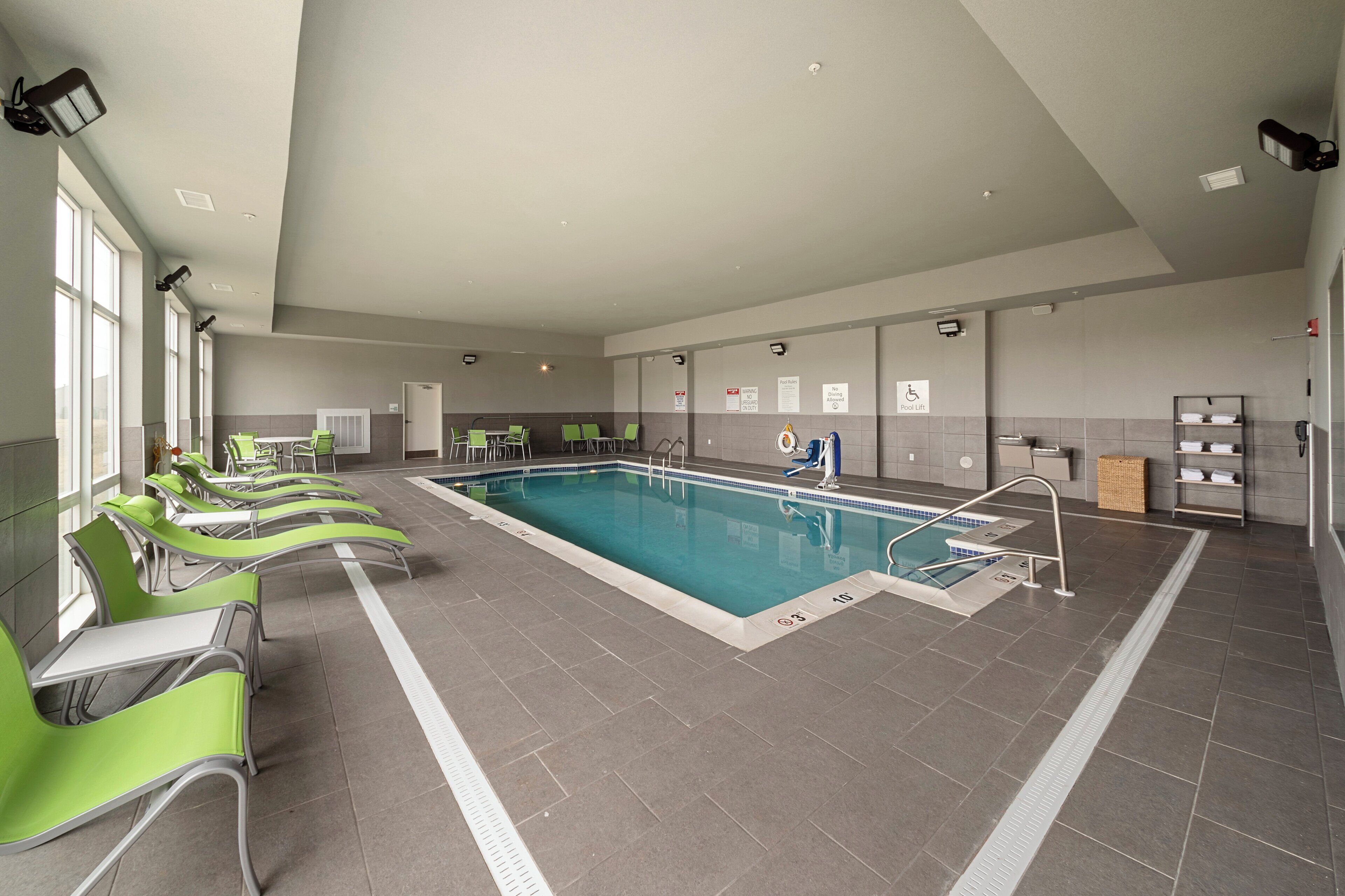 A swimming pool with a pool lift, green lounge chairs, and a tiled floor.
