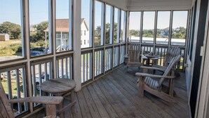 A covered porch with wooden chairs and a table, large windows offering views of the surrounding area. - Spartina 3 Bedroom Cottage by RedAwning (Buxton)