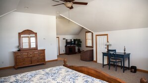 A bedroom with a large bed, a wooden dresser, a desk with a chair, a ceiling fan, and a view into another room. - Spartina 3 Bedroom Cottage by RedAwning (Buxton)