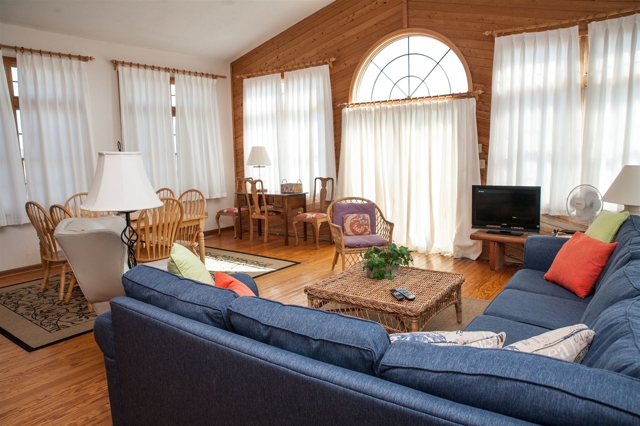 A cozy living room with a blue sofa, a dining table with chairs, a TV, and a large window with curtains.