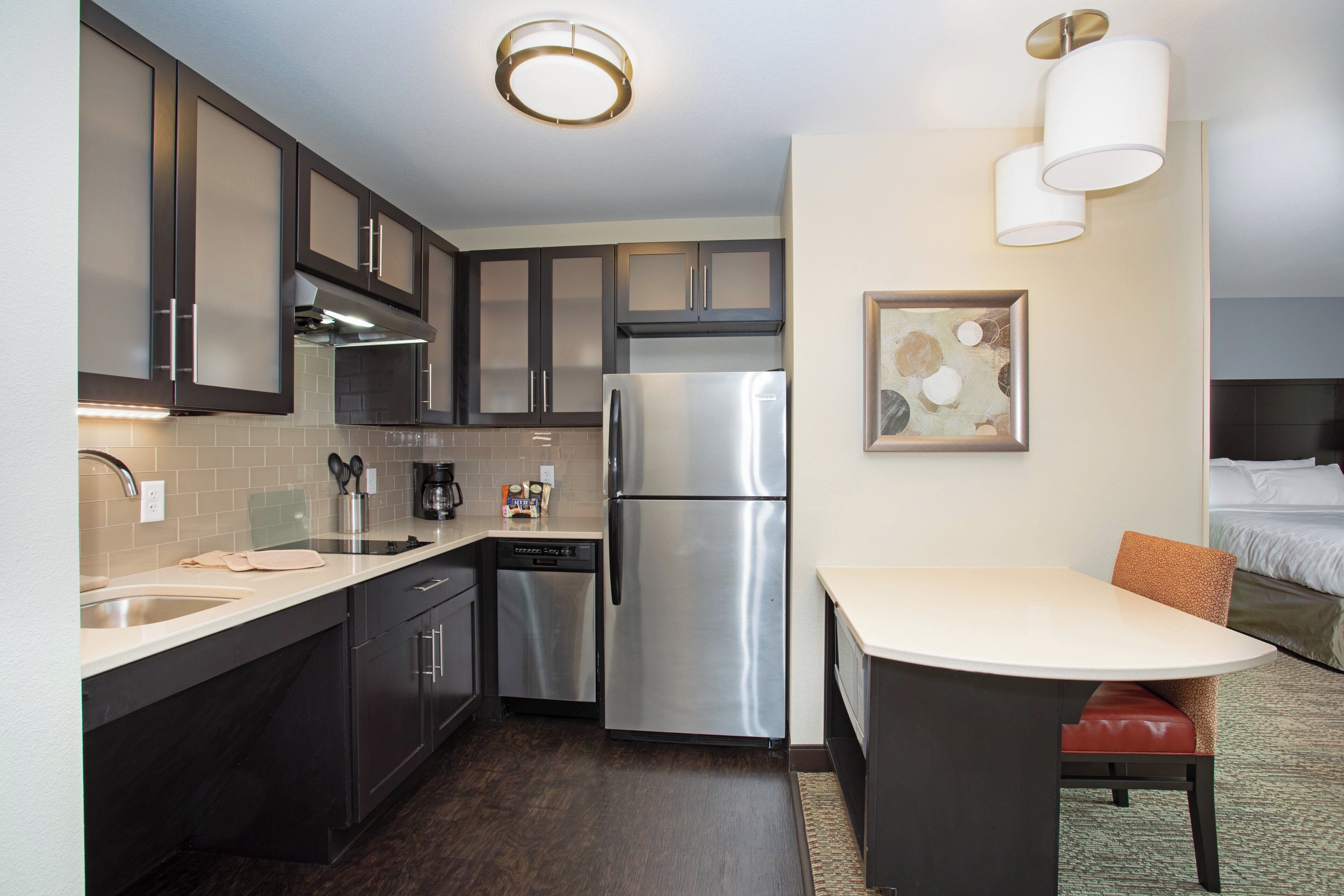 A compact hotel kitchen with a built-in dishwasher, a stainless steel refrigerator, and a dining area with a table and chair.