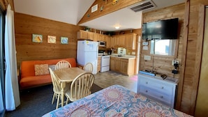 A compact kitchen with wooden cabinets, a refrigerator, and a microwave. There is a dining area with a table and chairs, and a sofa. A ceiling fan is visible above the kitchen. - 11 Mystical Sundaze Studio Bedroom Condo by RedAwning (Hatteras)