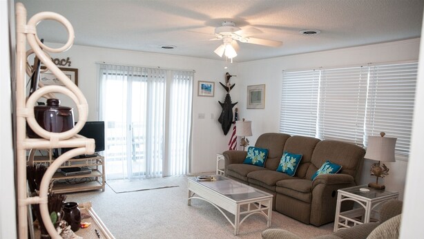 A living room with a sofa, coffee table, and a ceiling fan. There is a window with blinds, a door, and a wall-mounted decoration.