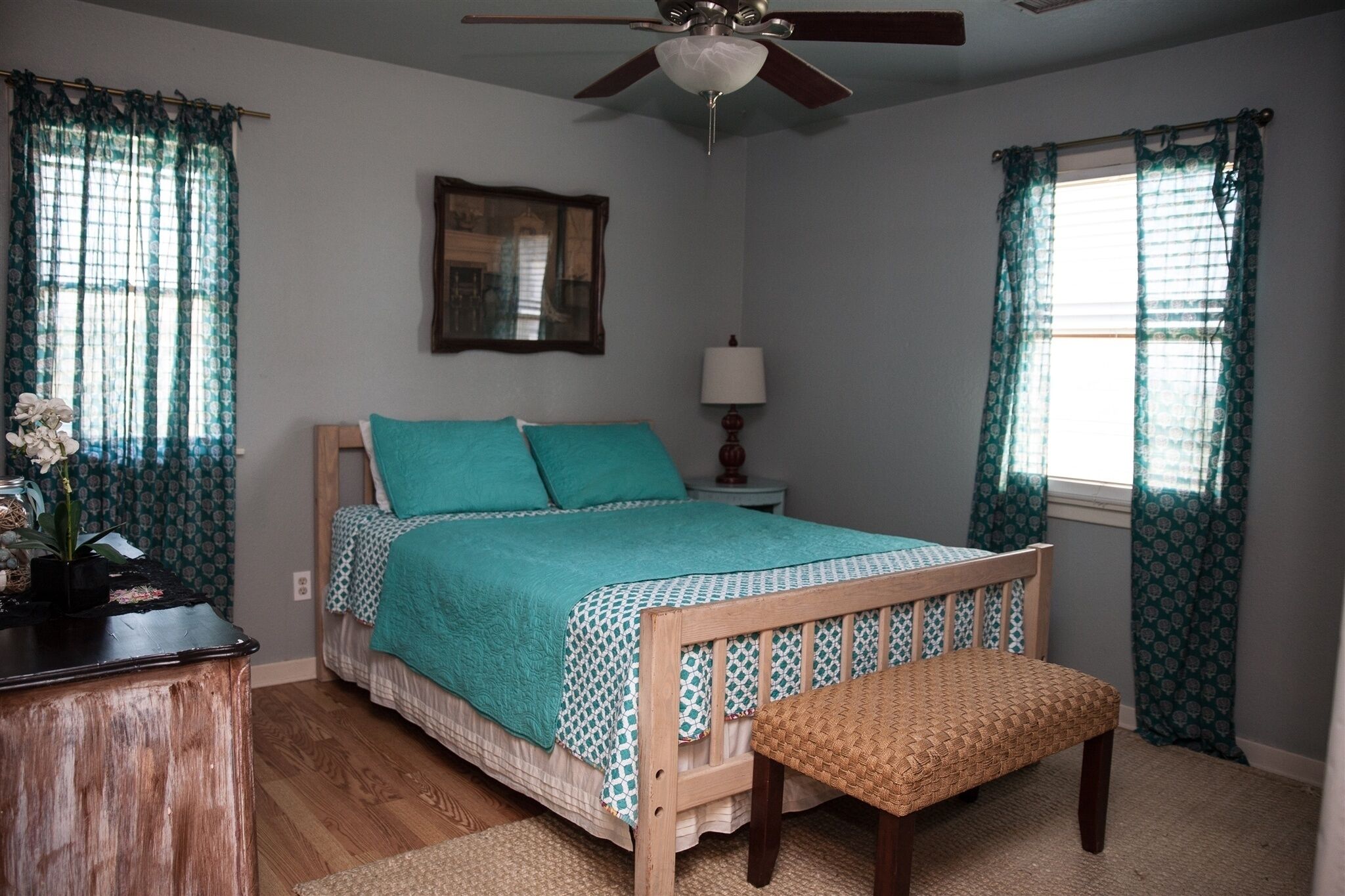 A bedroom with a bed, a wooden bench, a nightstand with a lamp, and a ceiling fan.
