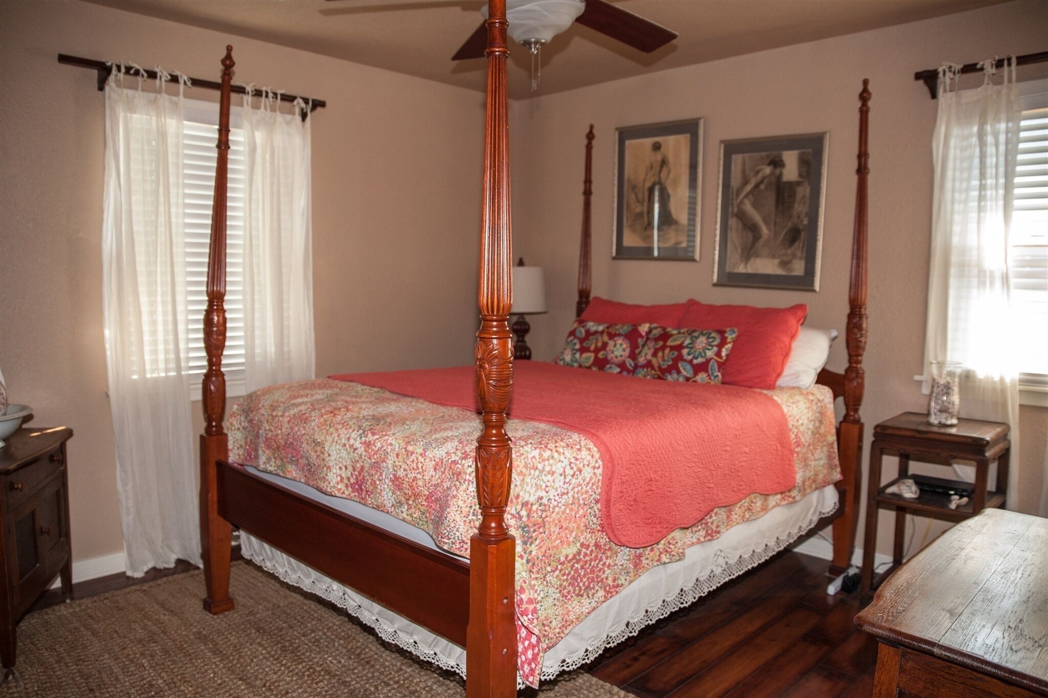 Una cama con dosel adornada con una colcha floral y almohadas rojas, una mesita de noche de madera con una lámpara y una pequeña mesa con un florero.