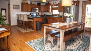 A kitchen with wooden cabinets, a stainless steel refrigerator, a dining table with benches, and a hanging light fixture. - Austin's Pointe 4 Bedroom Cottage by RedAwning (Hatteras)
