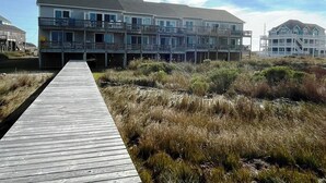 A multi-story beachfront building with balconies, a wooden boardwalk leading to it, and a grassy area in front.