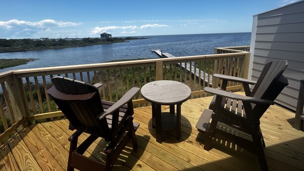 A wooden deck with two Adirondack chairs and a small round table, overlooking a body of water with a distant shoreline. - Sw 14 Shell Shack 1 Bedroom Condo by RedAwning (Hatteras)