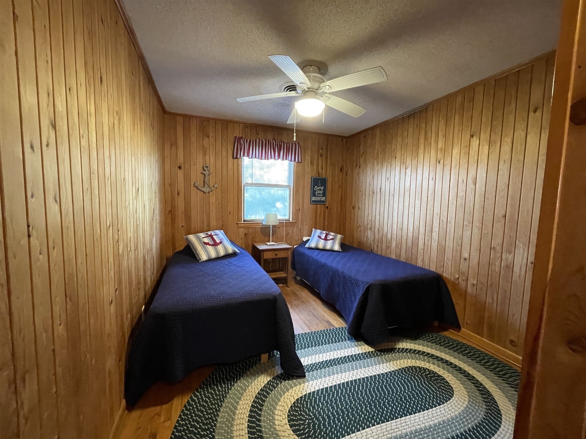Una habitación con paneles de madera, dos camas individuales con ropa de cama azul, un ventilador de techo, una ventana con cortinas y una alfombra en el suelo.